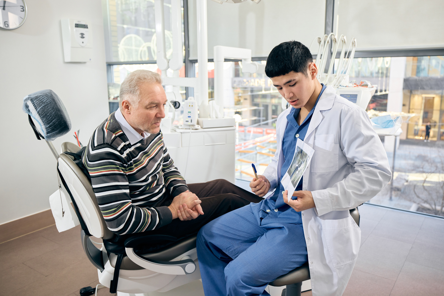 Dentist holding pen, shows x-ray to mature man, patient reclined in chair, explaining findings in bright, modern dental office setting. Concept of healthcare and pensioners, medicine.