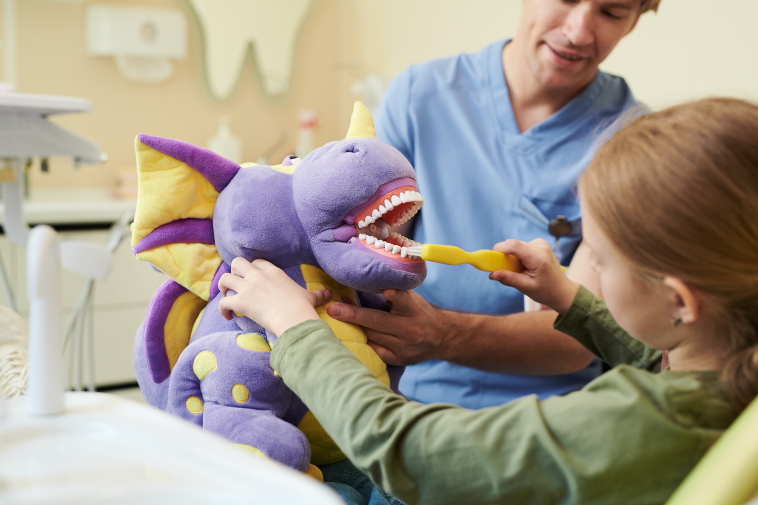 Caucasian girl child brushing teeth of purple dragon plush toy while young adult Caucasian male dentist supervising during dental education session in clinic setting
