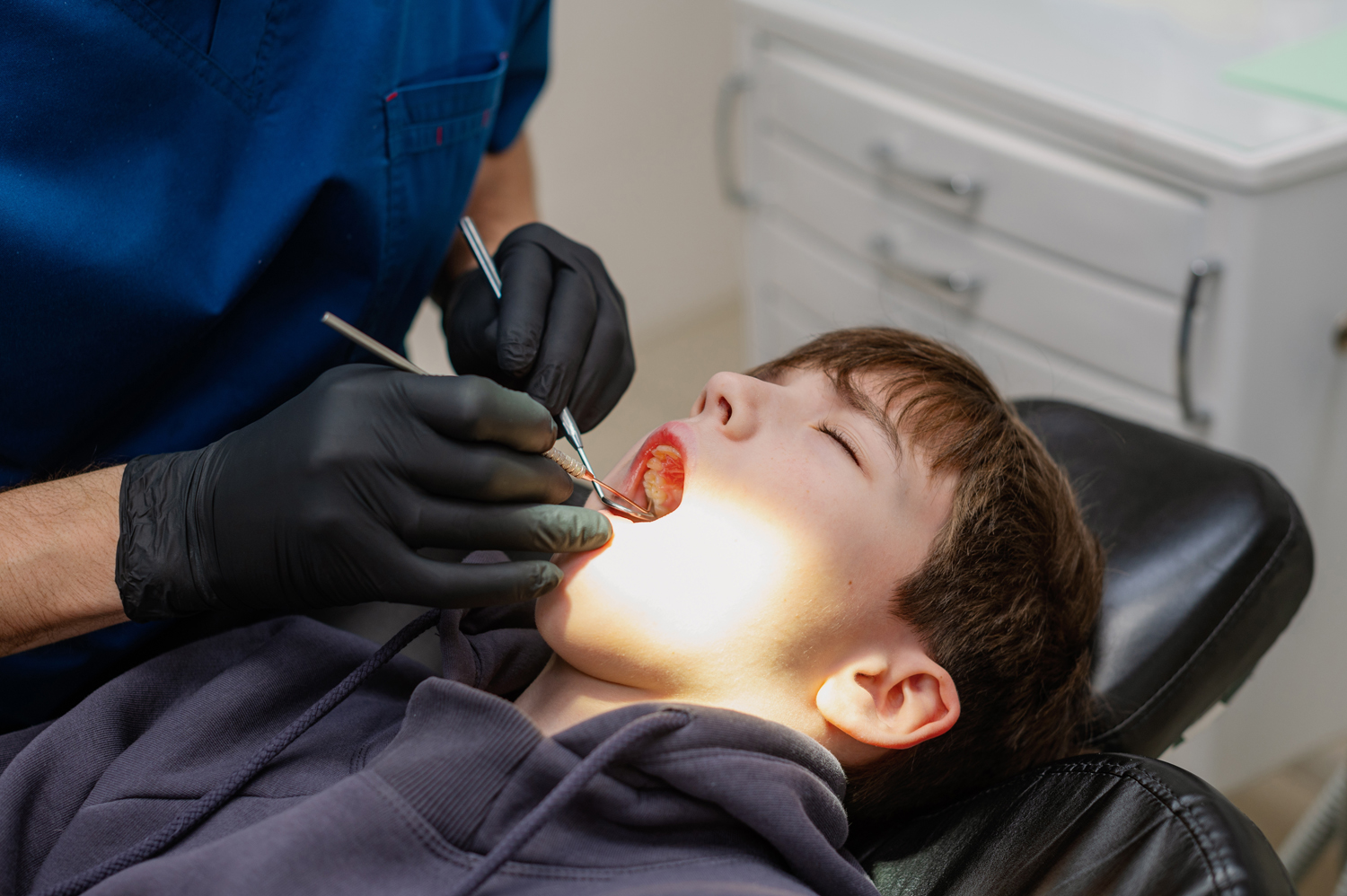 A dentist checks the teeth of a child who is sitting in an armchair with his mouth wide open. Routine dental check-up. Health